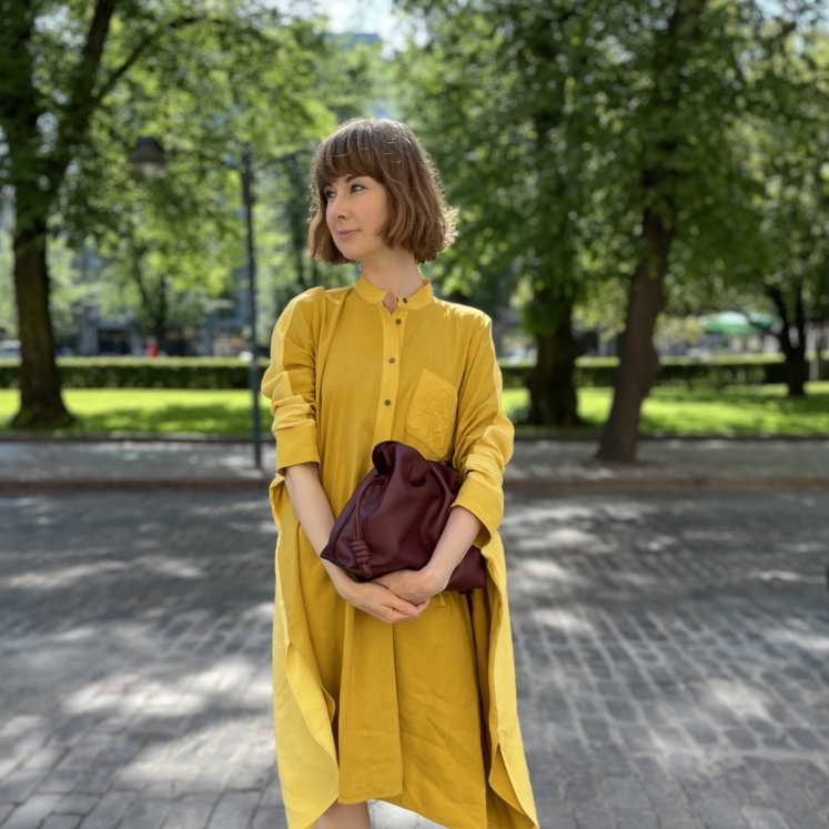nainen seisoo puistossa keltainen mekko yllään ja käsilaukku kädessään.