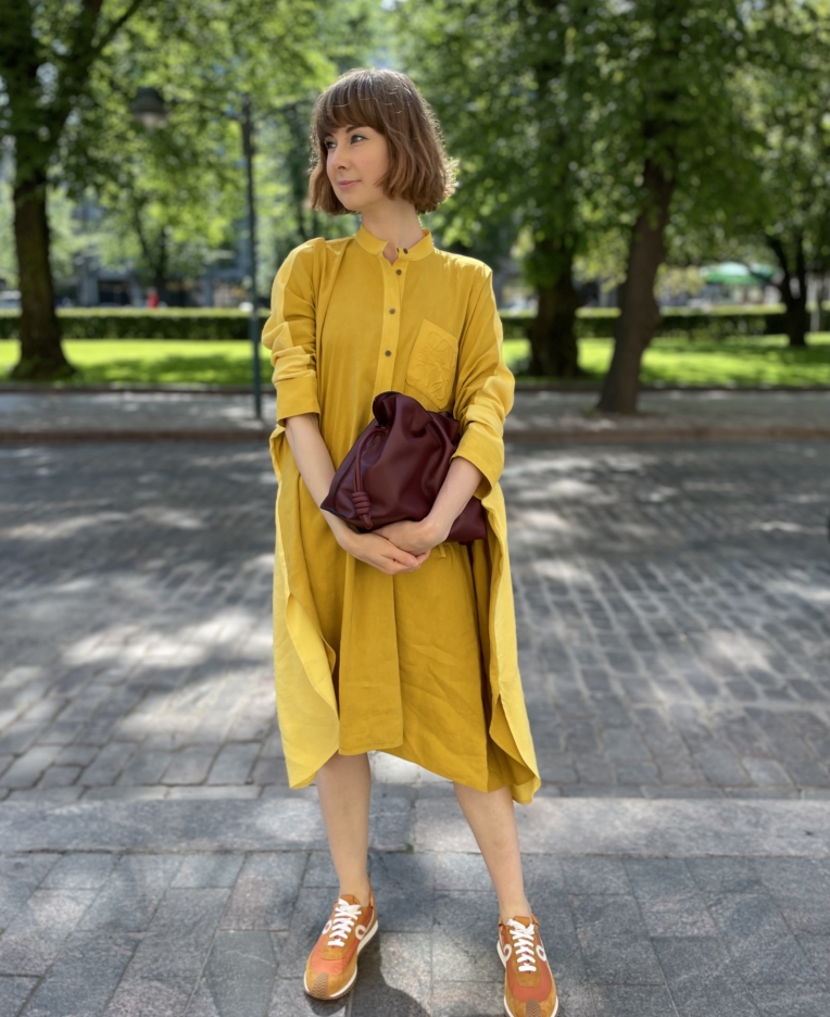 nainen seisoo puistossa keltainen mekko yllään ja käsilaukku kädessään.